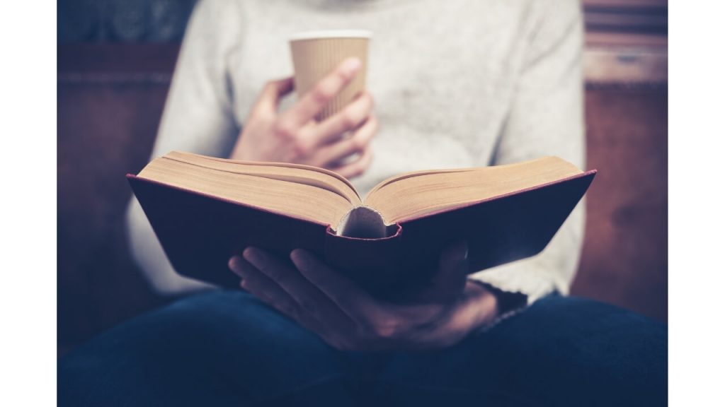An adult sitting and drinking a cup of coffee while reading a book