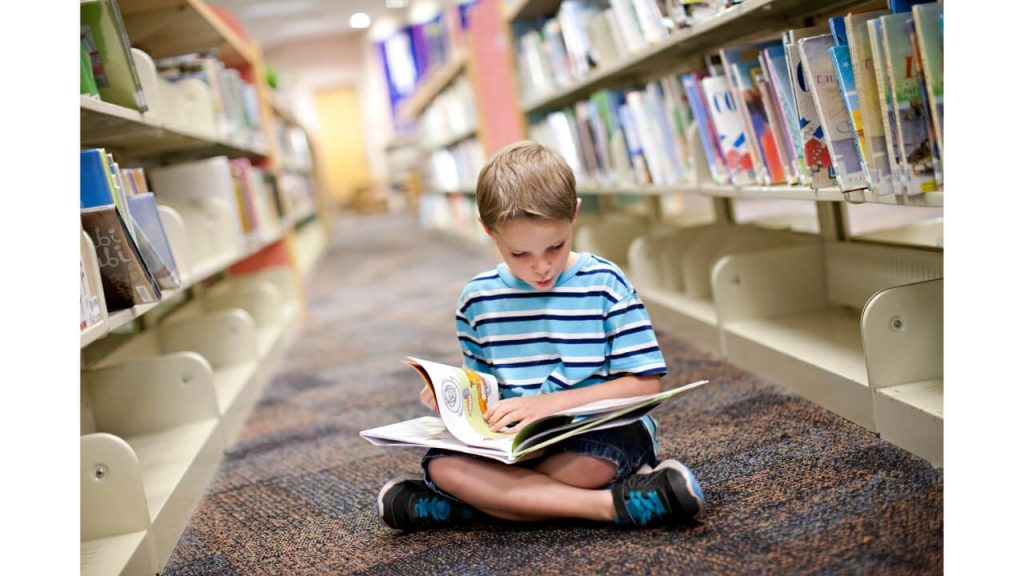 A small boy sitting in a library reading a book with stacks of book beside him