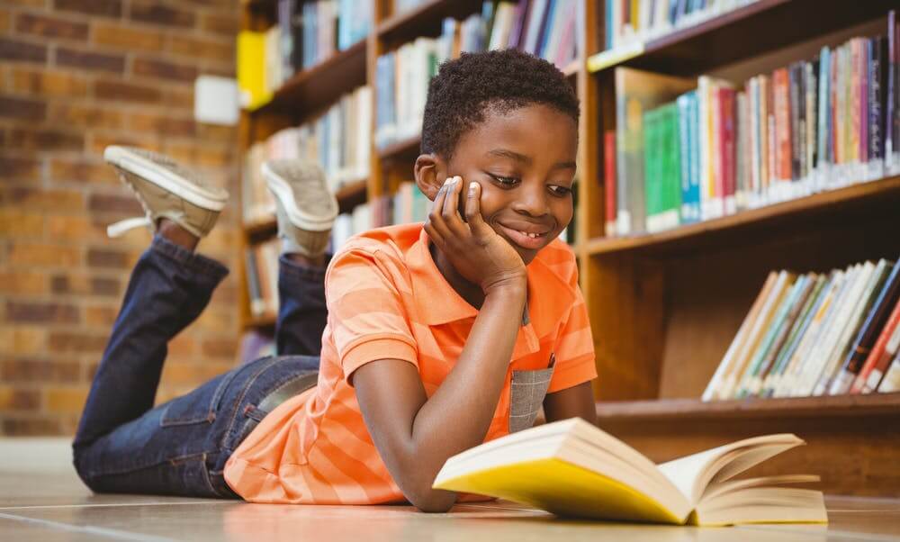 A young black boy reading a book in a library smiling laying on the floor on his stomach